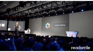 Audience watching a presentation on Referral Tracking. Large stage with multiple presenters and screens, including one displaying a woman and another showing a multi-colored gear logo.