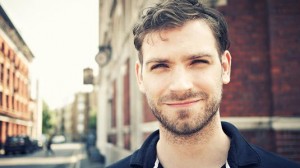Man with a short beard and dark hair stands on a city street looking slightly upward. Buildings line both sides of the street.