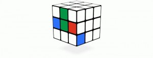 A Rubik's Cube sits angled on a white surface. The cube is mainly white, but one side has a row of blue, green, and red tiles. A shadow is visible directly below.